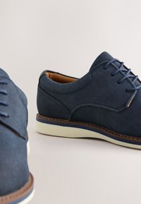 Paire de chaussures en daim bleu marine à lacets avec semelles blanches et bordure marron sur fond clair.
