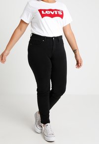 Jeans negros ajustados de cintura alta, combinados con una camiseta blanca que presenta un destacado logo rojo de Levi's, y zapatillas blancas.