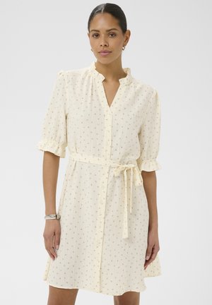 Kobieta w kremowej sukience zapinanej na guziki, z drobnymi kwiatowymi wzorami, falbanami na rękawach oraz paskiem zawiązanym w talii, stojąca na jednolitym tle.