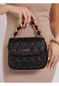 Sort, kviltet håndtaske med gyldne nitter og et flettet guld- og sort kædehåndtag. Har en frontklap med "GUESS EST 1981" logo.