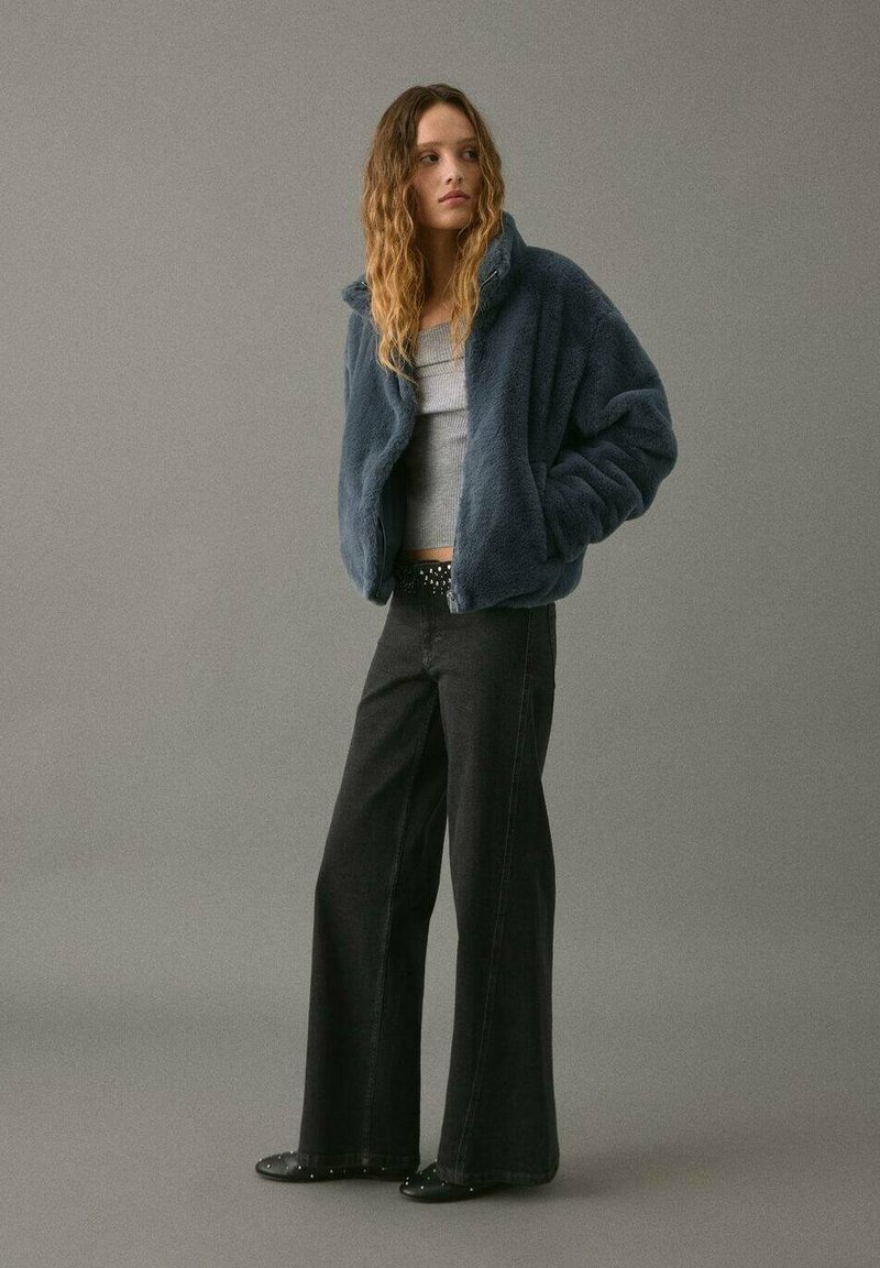 Žena s vlnitými vlasy nosí modrou umělou koženou bundu, šedý žebrovaný crop top, černé kalhoty s širokými nohavicemi, opasek s cvočky a černé boty s cvočky na šedém pozadí.