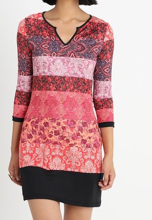 Femme portant une robe colorée à motifs avec des manches trois-quarts et un ourlet noir ajusté sur fond blanc.
