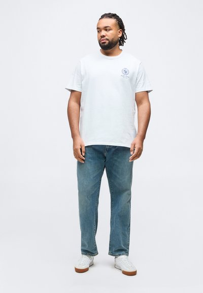 Άνδρας με κοτσίδες, που φοράει λευκό μπλουζάκι με στάμπα, μπλε τζιν και λευκά αθλητικά παπούτσια, στέκεται μπροστά σε ένα απλό ανοιχτό φόντο.