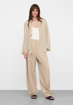 Femme portant un pantalon beige à jambes larges, un haut sans manches crème, un cardigan ouvert beige clair et des sandales noires à bout ouvert, debout devant un fond uni.