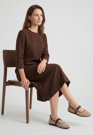 Frau sitzt auf einem Holzstuhl, trägt ein braunes Kleid mit langen Ärmeln und beige Mary Jane Schuhe mit dunklen Riemen vor einem schlichten Hintergrund.