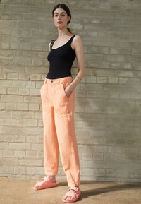 Top sin mangas negro ajustado, pantalones de pierna ancha en color naranja pastel con bolsillos frontales, y sandalias rosas con tiras ajustables sobre una superficie texturizada.
