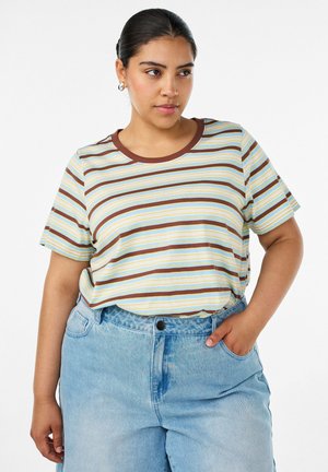 Kvinde iført en kortærmet, brun, gul og hvid stribet skjorte, stukket ned i lyseblå højtaljede jeans med den ene hånd i lommen.