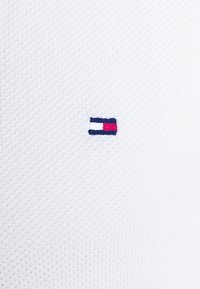 Petite broderie rectangulaire en bleu marine, blanc et rouge sur tissu blanc texturé, ressemblant à un logo de marque sur des vêtements.