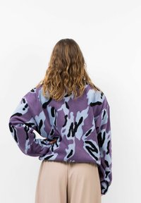 Fleece-Pullover im lila-blauen Camouflage-Muster mit schwarzen Akzenten, mit lockerer Passform, überschnittenen Schultern und weicher Textur.