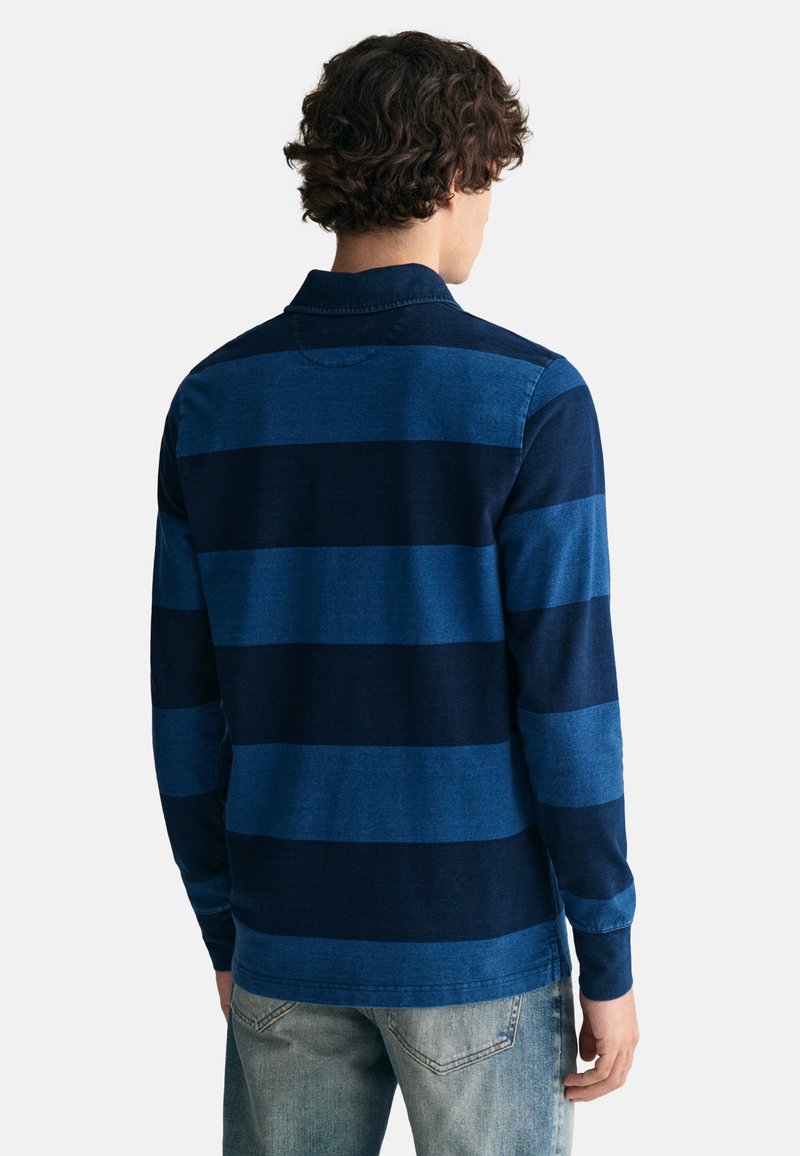 Uomo con capelli ricci che indossa una camicia a righe blu navy e blu a maniche lunghe e jeans azzurri, di spalle su sfondo bianco.