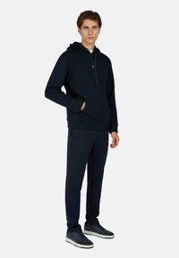 Felpa con cappuccio navy, dotata di tasca a marsupio, chiusura con cordino e polsini a coste, abbinata a pantaloni della tuta coordinati e sneaker scure.