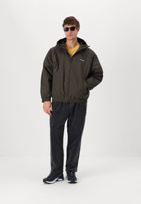 Veste légère vert olive foncé avec capuche, fermeture éclair à l'avant et poignets élastiques. Assortie à un pantalon en velours côtelé noir et des baskets noires.