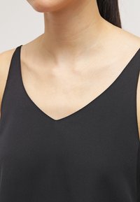 Débardeur noir sans manches avec un design en V, fabriqué en tissu lisse, présentant une coupe ajustée et un minimalisme au niveau de l'encolure.