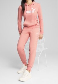 Sudadera rosa con logo blanco de Puma y bolsillo tipo canguro, combinada con pantalones de chándal rosa a juego con logo, y zapatillas deportivas blancas.