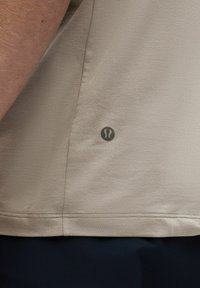 Beige színű sportpóló strukturált anyagból, kis fekete logóval az alsó oldalon. A kivágás egyenes, látható varrási részletekkel.