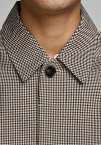 Detailní záběr na osobu, která nosí hnědo-bílý kabát s kostkovaným vzorem a velkým černým knoflíkem zapnutým na límci.