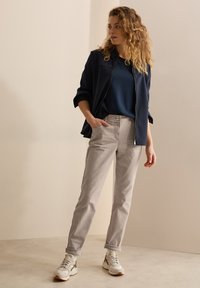 Chaqueta de cremallera azul marino sobre una camiseta azul, combinada con pantalones gris claro y zapatillas blancas con suela beige. El modelo está de pie con una mano en el bolsillo.