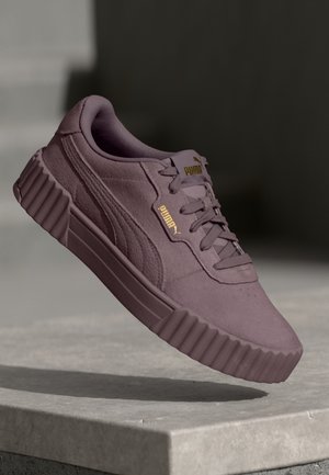 Baskets en daim violet Puma avec semelle épaisse à rainures et logos Puma dorés, en angle au-dessus d'une dalle de béton sur un fond sombre.