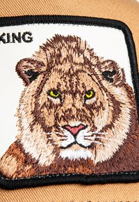 Parche bordado con la cara de un león en tonos de marrón y crema, contorneado en negro sobre un fondo beige. El texto arriba dice "REY."