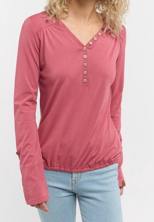 Femme portant un chemisier rose poudré à manches longues avec un col en V, six boutons beige et un jean bleu clair sur un fond blanc uni.