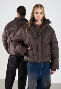 Două persoane tinere purtând jachete puf dintr-un maron închis și blugi largi, una întorcându-se cu fața spre cameră și cealaltă cu spatele.