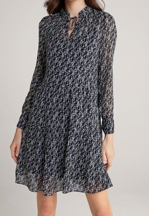 Donna che indossa un vestito a maniche lunghe con motivo bianco e nero, scollo a occhiello con fiocco e gonna svasata.