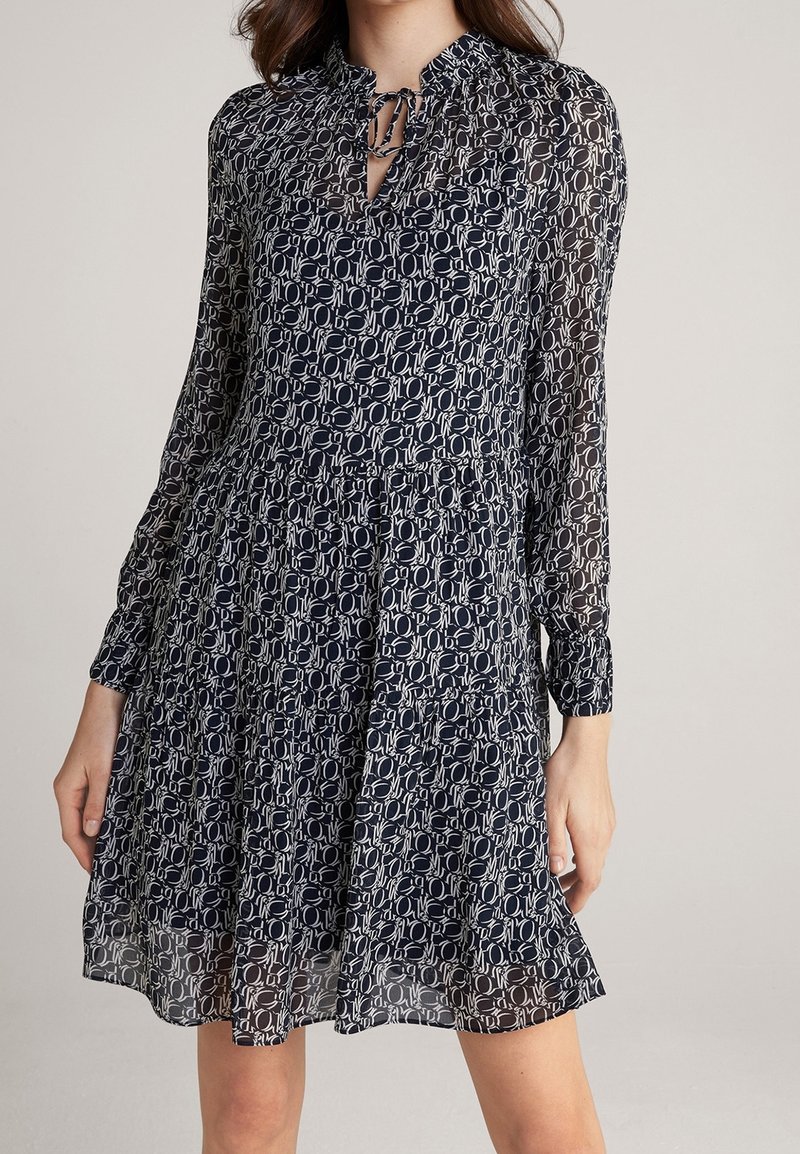 Femme portant une robe à manches longues à motifs noirs et blancs, avec une encolure à clé nouée et une jupe évasée.