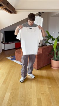 Jovem vestindo uma camisa cinza e calças cargo, segurando uma t-shirt branca, dentro de uma sala de estar moderna com pisos de madeira e plantas.