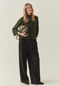 Zaļš adīts džemperis ar augstu apkakli, savienots ar melniem platām biksēm. Tur rokās strukturētu olīvu krāsas rokassomu, melnas kurpes.