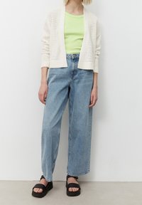 Cardigan crema a maglia sopra una camicia verde chiaro, abbinato a jeans blu a gamba larga e sandali con piattaforma neri. Abbigliamento strutturato e dal fitting morbido.