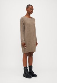 Vestido de malha castanho claro, com um corte descontraído, ombros caídos e punhos canelados. Combinado com botins pretos, acabamento texturizado.