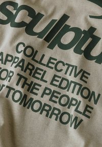 Gros plan sur un tissu beige avec un texte vert foncé : "ÉDITION VÊTEMENTS COLLECTIF DE SCULPTURE POUR LE PEUPLE DE DEMAIN."