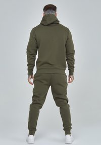 Olivgrüner Hoodie- und Jogginganzug aus weichem Stoff. Verfügt über eine Kapuze, eine lässige Passform und gerippte Bündchen, kombiniert mit weißen Sneakers.