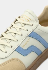 A sportcipő világos krémszínű bőr felsőrésszel, kék kiemelésekkel, bézs színű velúr orrkialakítással és texturált gumi talppal rendelkezik. Arany logó részlet is megtalálható.