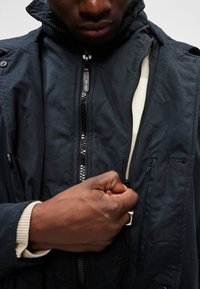Casaco azul marinho com gola alta e múltiplos bolsos, apresentando um tecido sólido, fecho de correr suave e punhos canelados. Detalhe de material texturizado visível.