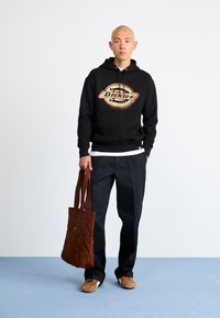 Μαύρο hoodie με λογότυπο "Dickies", μαύρο παντελόνι, καφέ καπιτονέ τσάντα ώμου και μπεζ αθλητικά παπούτσια. Απλό φόντο.