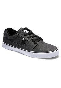 Las zapatillas de skate de lona negra presentan un diseño de corte bajo, suela de goma blanca, cordones negros y un pequeño logo en el lateral. Se aprecia un detalle de superficie texturizada.
