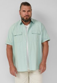 Hellblaues kurzärmliges Hemd aus strukturiertem Stoff, mit zwei Brusttaschen und weißen Knöpfen. Über einem weißen T-Shirt getragen.