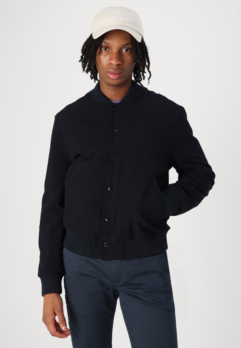 Giacca bomber lavorata a maglia di colore blu navy con orlo a coste, chiusura con bottoni e tasche laterali. Abbinata a pantaloni blu scuro e un cappellino chiaro.