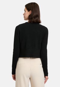 Pull court noir avec une finition texturée, col rond et manches longues. Porté avec un pantalon clair, vu de dos.