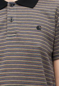 Κοντινό πλάνο ενός ατόμου που φοράει πολύχρωμη πόλο μπλούζα με οριζόντιες ρίγες, μαύρο γιακά και κουμπιά, με μαύρο κεντημένο λογότυπο στο στήθος.