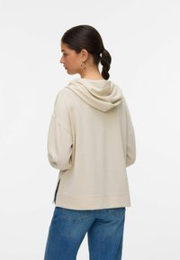 Beige mikina s měkkou strukturou, oversized střih a bočními rozparky. Nosená s volnými modrými džíny s jemnými detaily prošití.
