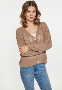 Bruine gebreide cardigan met een V-hals, open geweven patroon en twee knopen. Gecombineerd met lichtblauwe jeans, die een ontspannen pasvorm behouden.
