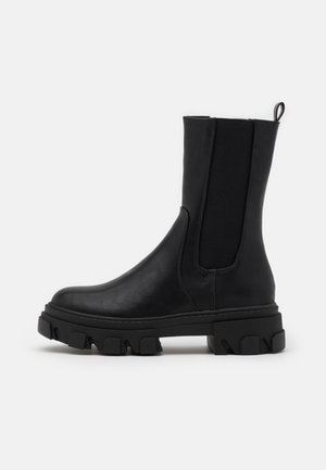 Musta nahkainen mid-calf Chelsea-saapas paksulla nastapohjalla, joustavilla sivupaneeleilla ja vetotapilla yksivärisellä taustalla.
