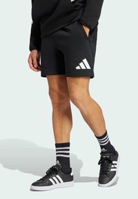 I pantaloni sportivi neri presentano una superficie testurizzata e un logo bianco a tre strisce. Indossati con calze nere e sneakers nere con accenti bianchi.