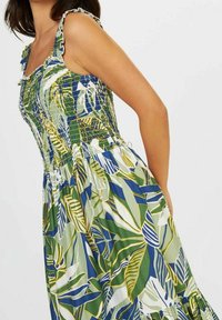 Robe fleurie avec corsage froncé, motif de feuilles vertes et bleues, bretelles à volants et ourlet de jupe évasé. Tissu léger.