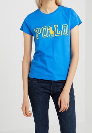 Femme portant un t-shirt bleu vif à manches courtes avec un texte jaune "POLO" et un graphisme de joueur de polo à cheval, associé à un jean bleu foncé.