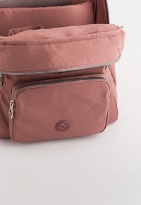 Mochila rosa polvorienta con un compartimento principal abierto y un bolsillo frontal cerrado con cremallera, sobre un fondo claro y liso.