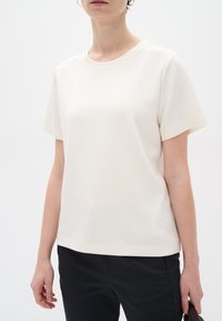 Cream-colored short-sleeve top with a rounded neckline, smooth texture, and subtle branding at the hem. Paired with dark pants.