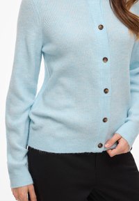 Ljusblå kofta i mjukt stickat tyg med knäppning fram, ribbad struktur och en kort fåll med lätt luddig yta.